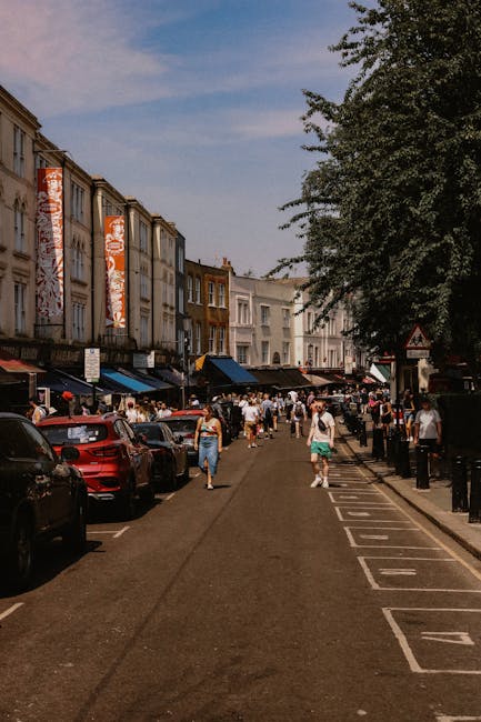 A busy street scene in Portobello Road, North Kensington, showing pedestrians walking along the pavement and a row of parked cars on the left side. On the right, there is a designated loading area wit