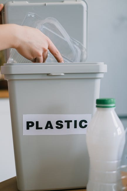 Close-up of a grey plastic waste bin labeled 'PLASTIC' in bold black letters, with the bin lid open and a person's hand placing a clear plastic container inside. In front of the bin, there is a partia
